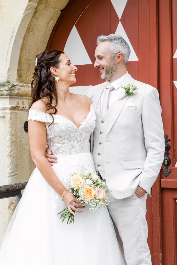 Nahaufnahme von Braut und Bräutigam in heller Hochzeitsmode, die sich vor einer roten Holztür liebevoll in die Augen sehen.