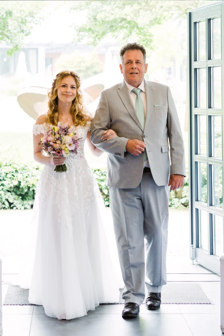Die Braut schreitet bei ihrer Landhotel Voshövel Hochzeit gemeinsam mit ihrem Vater in einem eleganten grauen Anzug in den Trausaal, während sie einen romantischen Blumenstrauß trägt.
