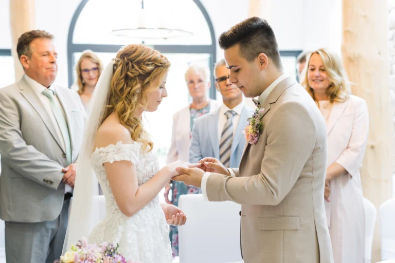 Nahaufnahme der Hände des Bräutigams, während er seiner Braut den Ehering ansteckt – ein bedeutungsvoller Moment der Trauung.