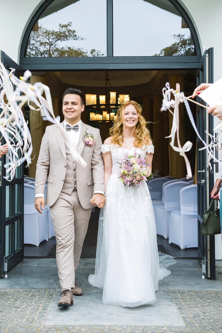 Braut und Bräutigam verlassen fröhlich das Confideum Voshövel, umgeben von jubelnden Gästen mit Wedding Wands.