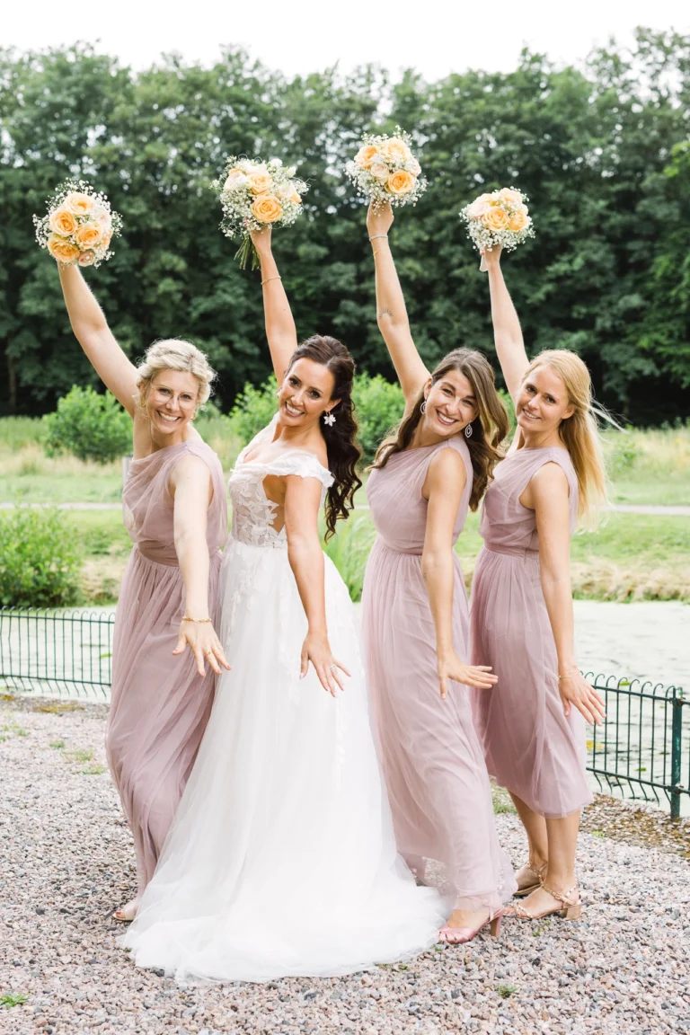 Braut und drei Brautjungfern in rosé Kleidern mit Blumensträußen vor grünem Park und Wasserschloss Raesfeld.