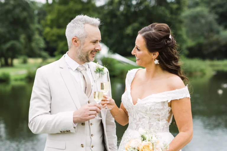 Braut und Bräutigam stoßen lächelnd mit Sektgläsern am Wassergraben von Schloss Raesfeld an – Hochzeitsfotograf Schloss Raesfeld.
