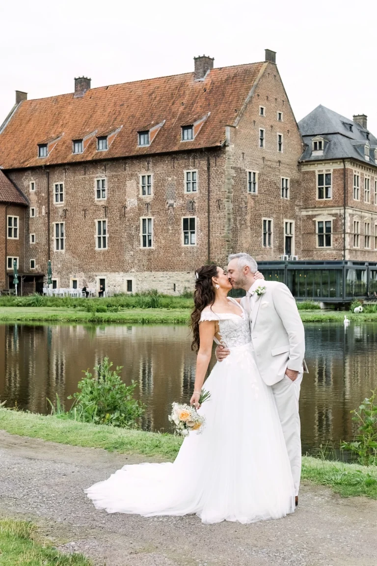 Braut und Bräutigam küssen sich vor dem Wasserschloss Raesfeld am spiegelnden Schlossgraben.