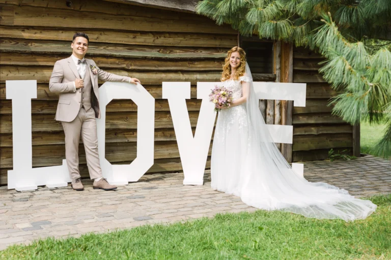 Braut und Bräutigam stehen neben riesigen LOVE-Buchstaben bei ihrer Landhotel Voshövel Hochzeit – ein romantischer Hingucker vor rustikaler Holzwand.