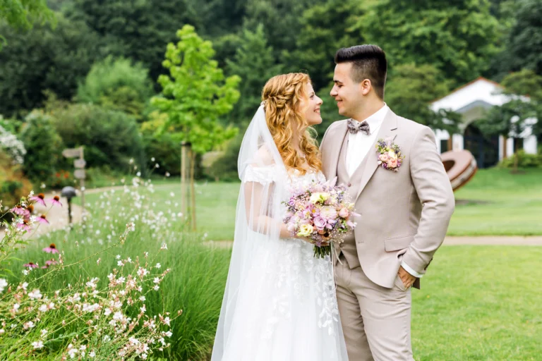 Romantisches Brautpaar bei ihrer Landhotel Voshövel Hochzeit – der Bräutigam im beigen Anzug mit Fliege, die Braut mit Schleier und Blumenstrauß, beide blicken sich verliebt an.