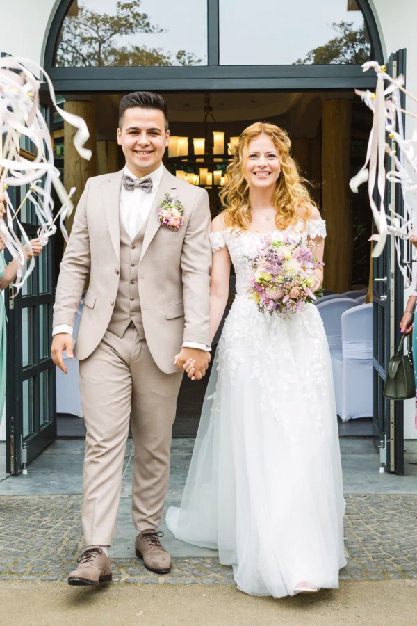 Braut und Bräutigam verlassen fröhlich das Confideum Voshövel, umgeben von jubelnden Gästen mit Wedding Wands.