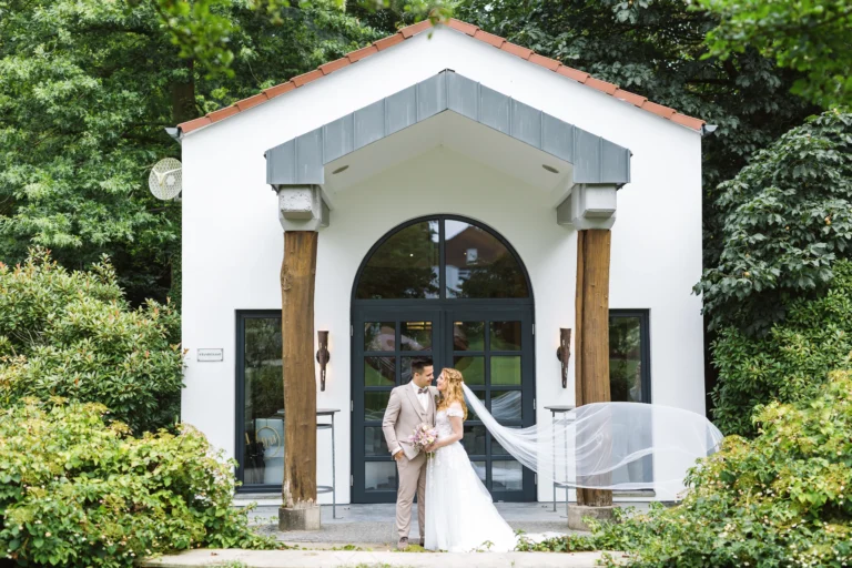 Romantisches Brautpaar steht vor dem Confideum Voshövel, umgeben von grüner Natur. Der Schleier der Braut weht im Wind und verleiht der Szene eine märchenhafte Atmosphäre.