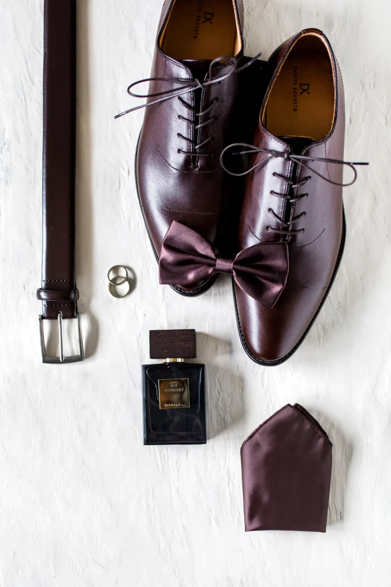 Bräutigam-Accessoires für das Getting Ready: Dunkle Lederschuhe, Fliege, Eheringe und Parfum stilvoll arrangiert.
